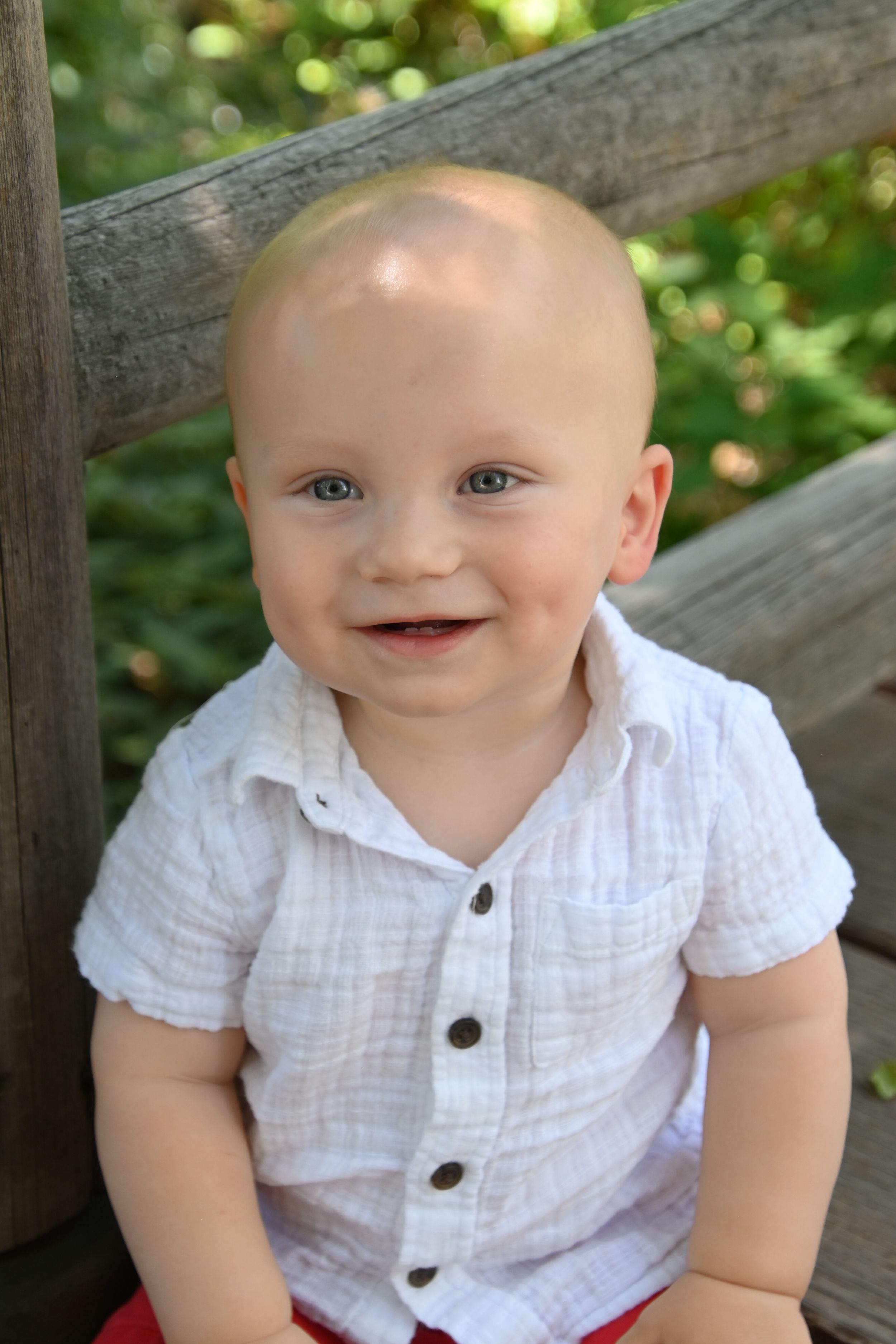 Happy baby wearing a white button-up shirt sits on wooden bench outdoors on sunny day.
