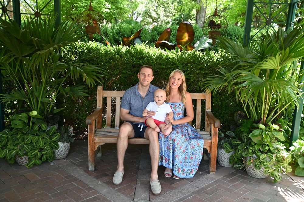 A couple sits together on a wooden bench surrounded by lush tropical plants and greenery in a garden setting.