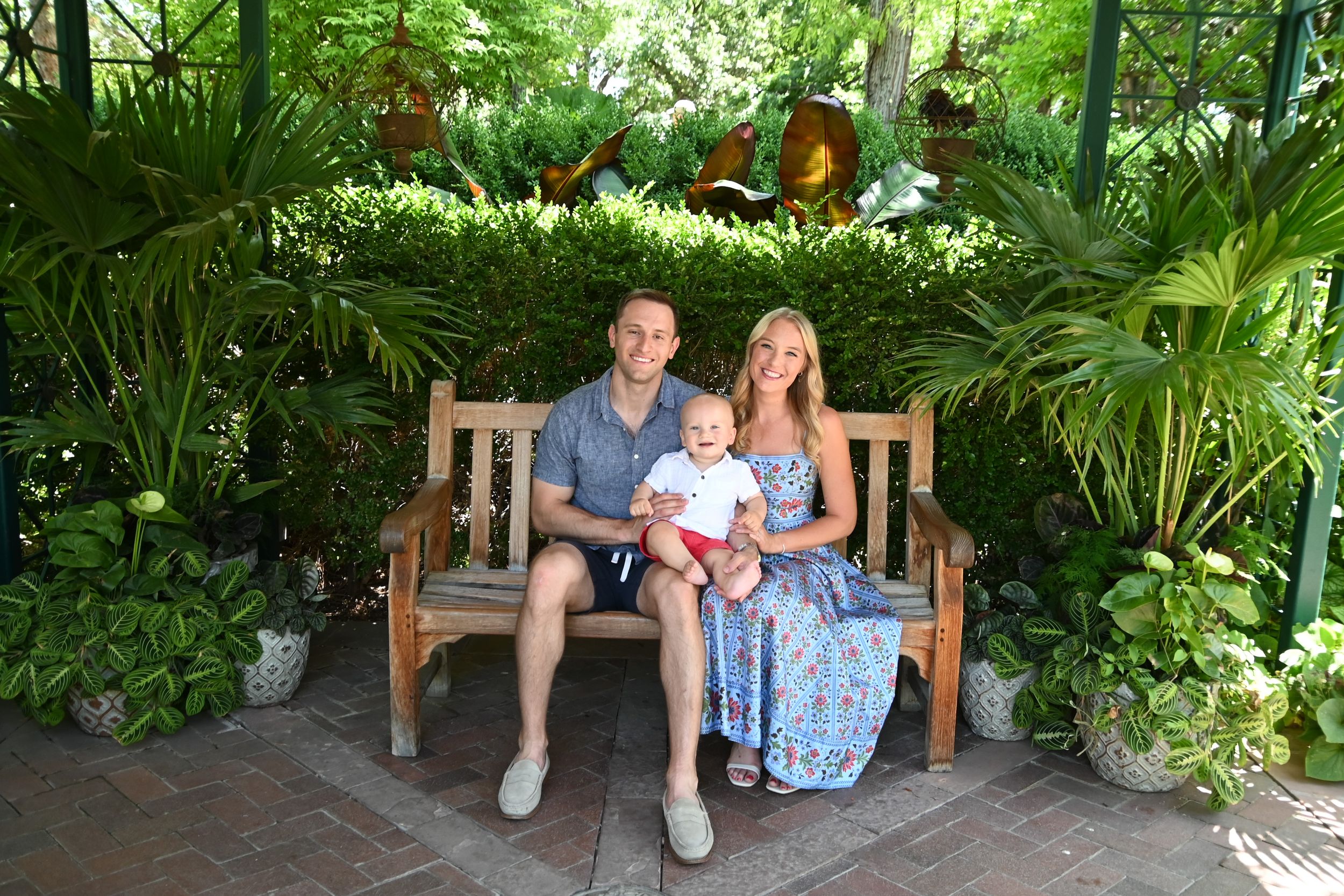 A couple sits together on a wooden bench surrounded by lush tropical plants and greenery in a garden setting.
