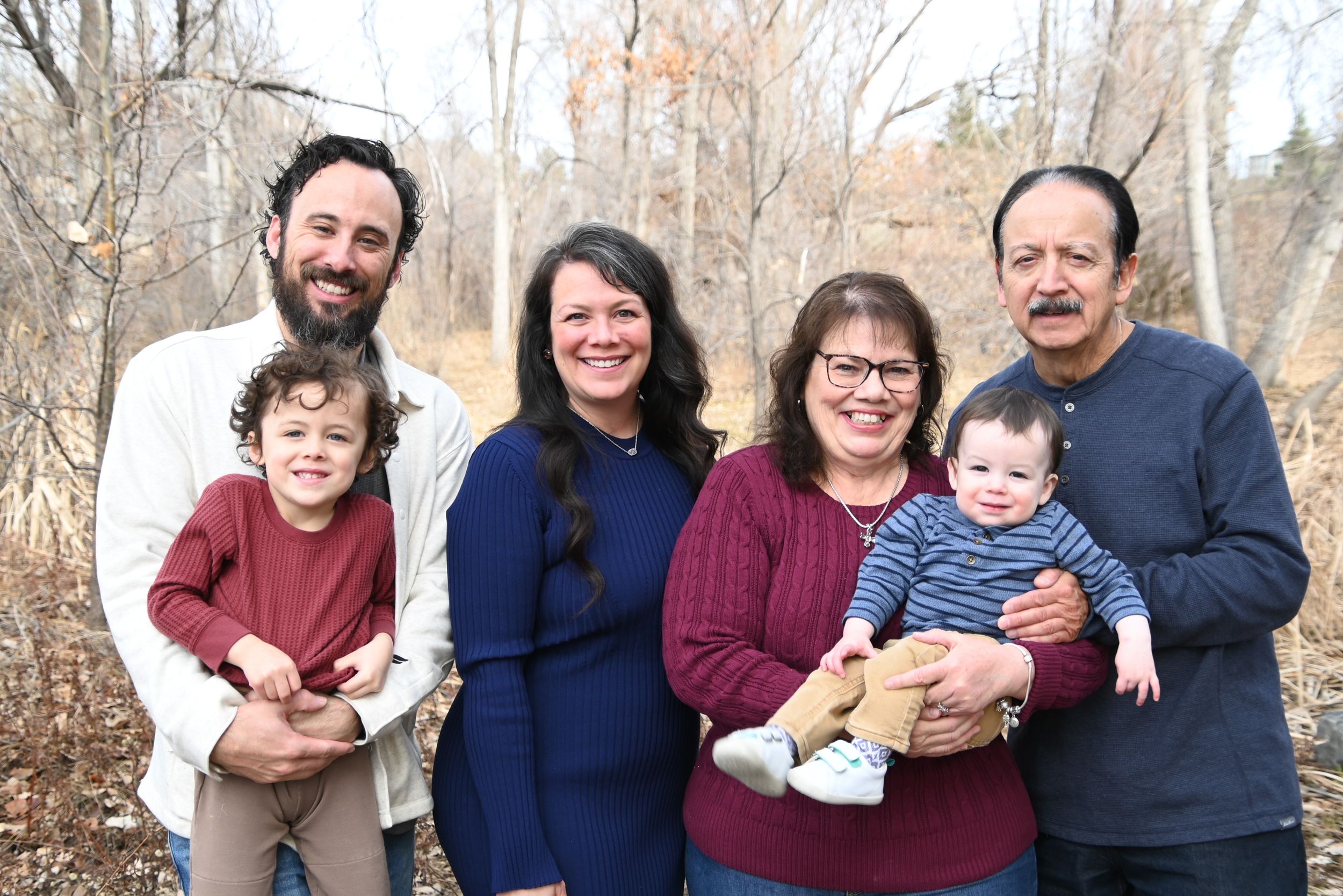 A multi-generational family group poses together outdoors on a fall day with bare trees in the background.