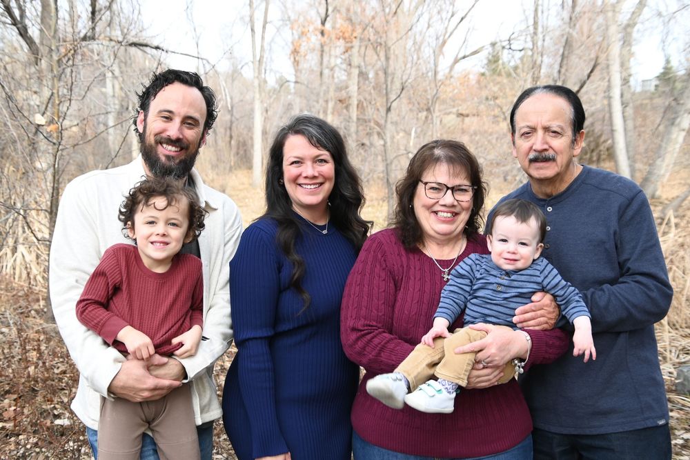 A multi-generational family group poses together outdoors on a fall day with bare trees in the background.