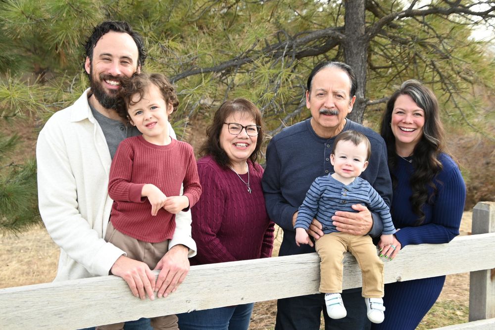 Multi-generational family group poses together outdoors against a backdrop of pine trees and wooden fence.
