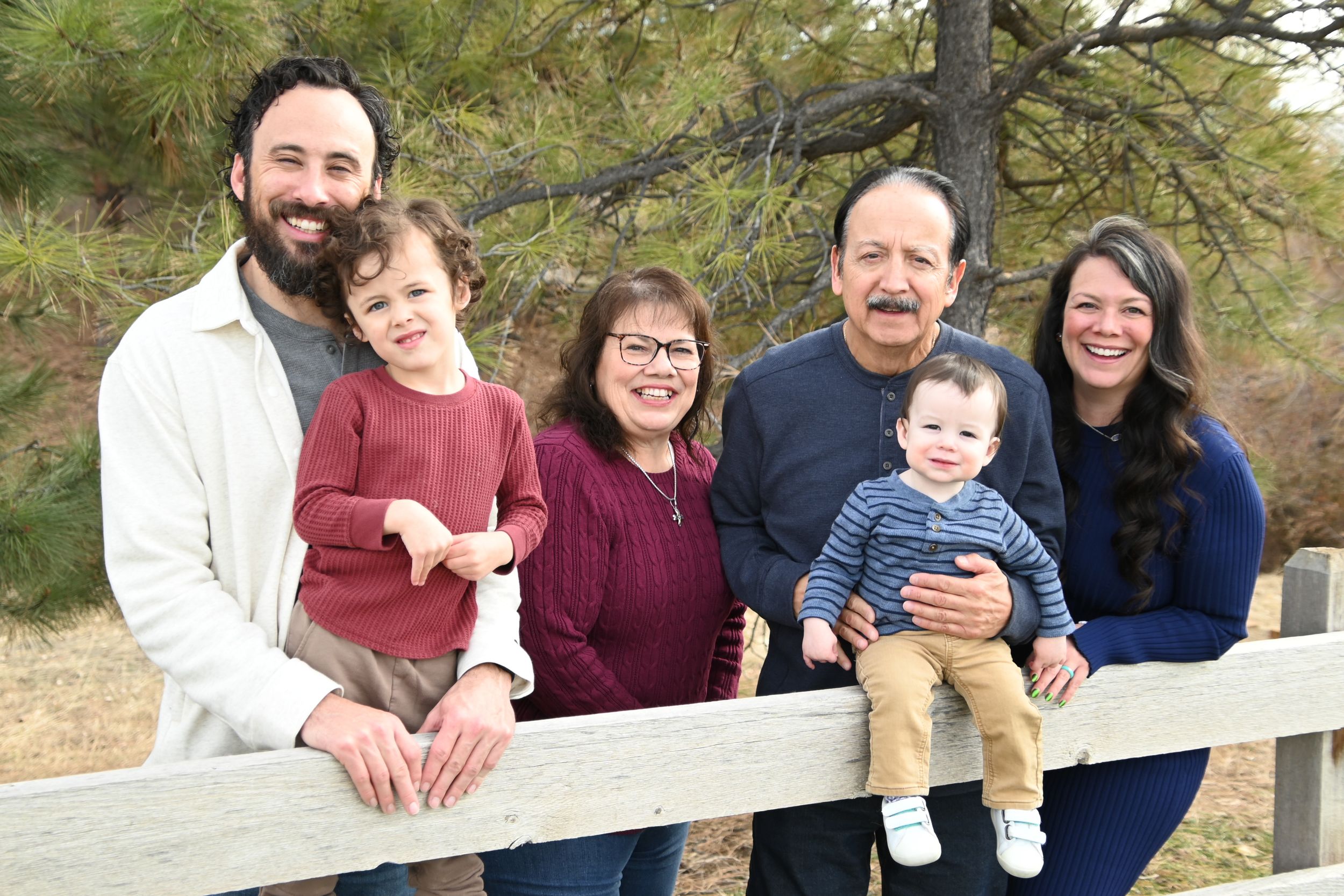Multi-generational family group poses together outdoors against a backdrop of pine trees and wooden fence.