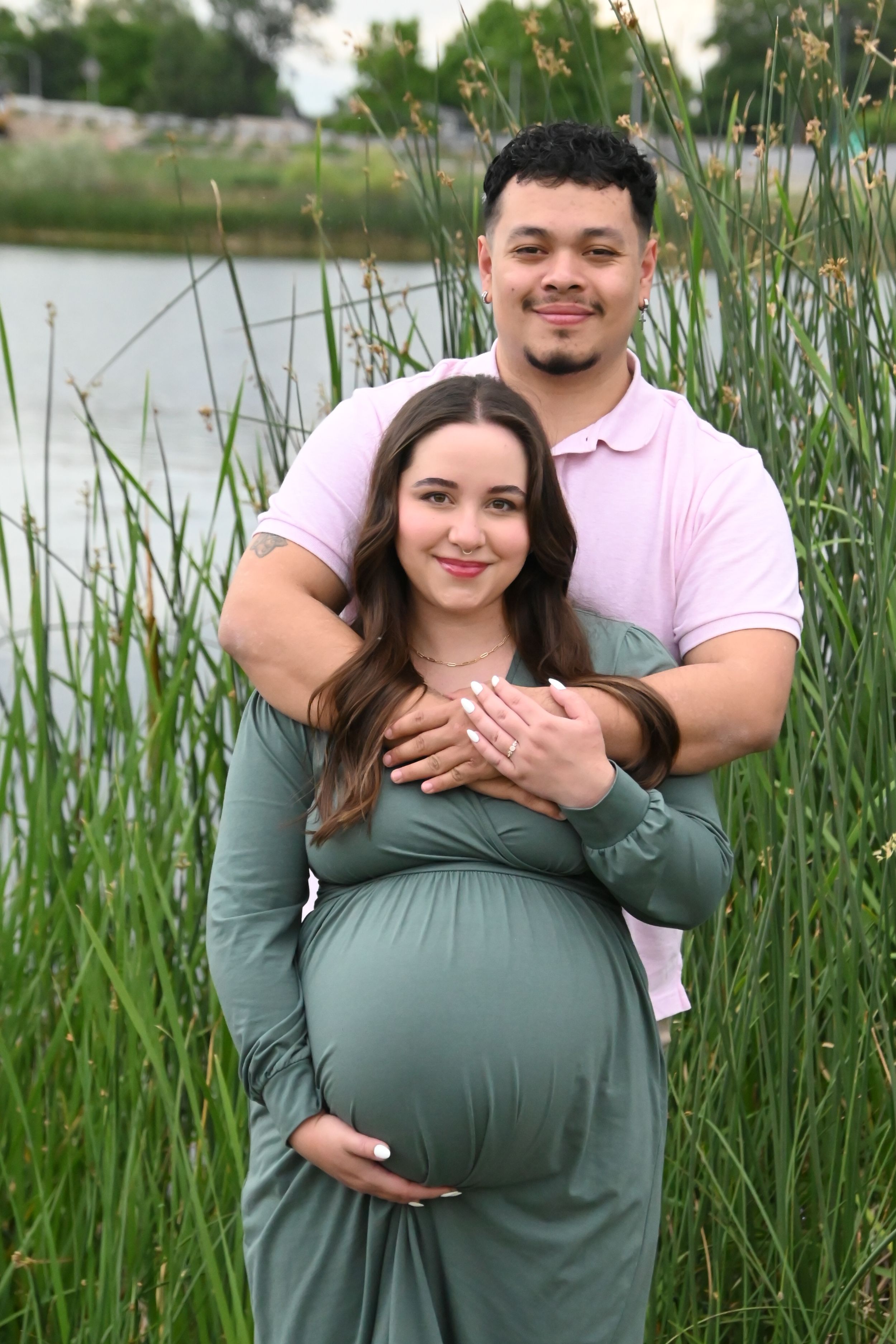 Expectant couple embraces in a peaceful outdoor setting with tall grass and pond in the background.