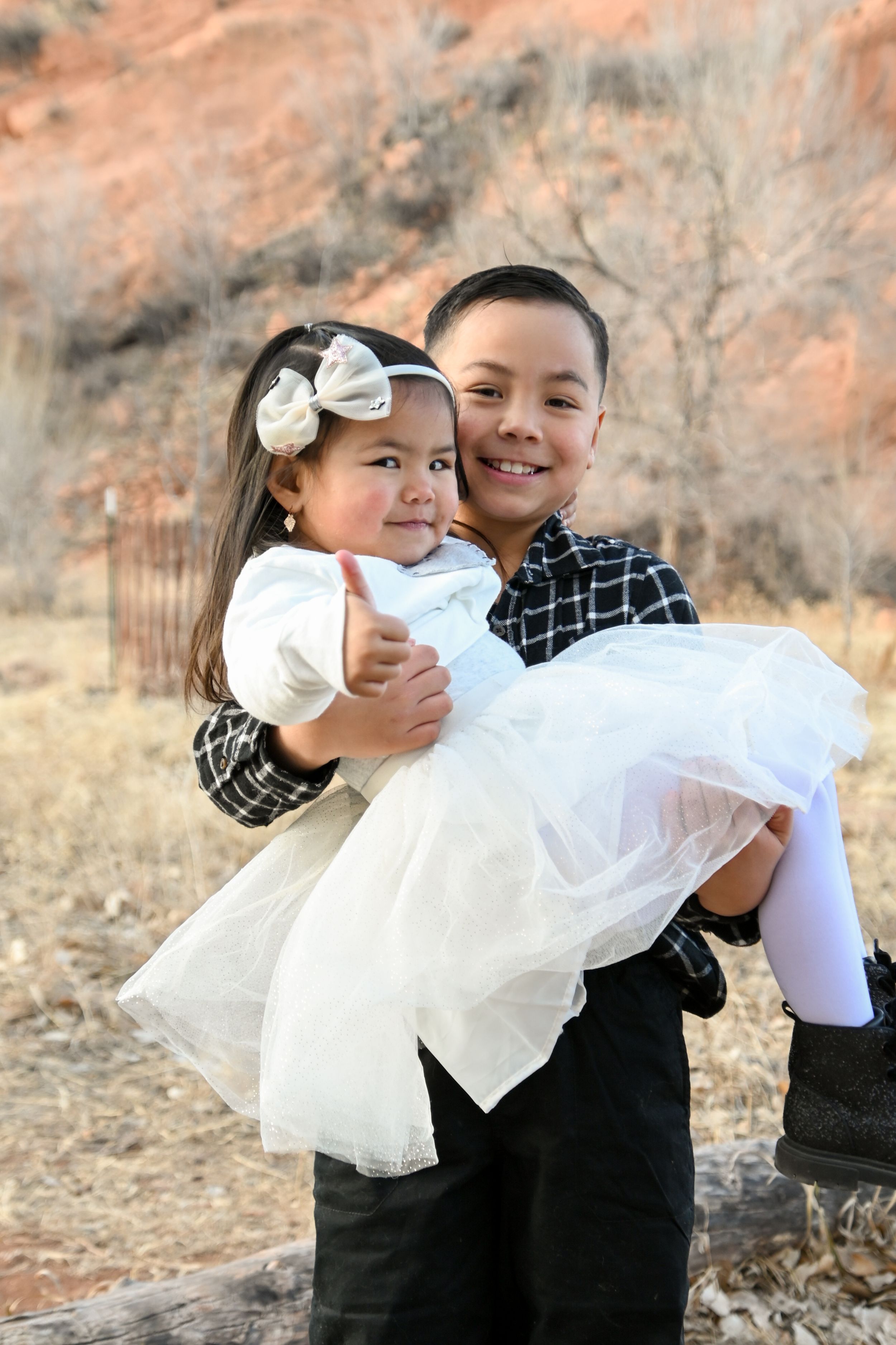 Two children in formal wear embracing outdoors against red rock background, with white dress and plaid shirt.
