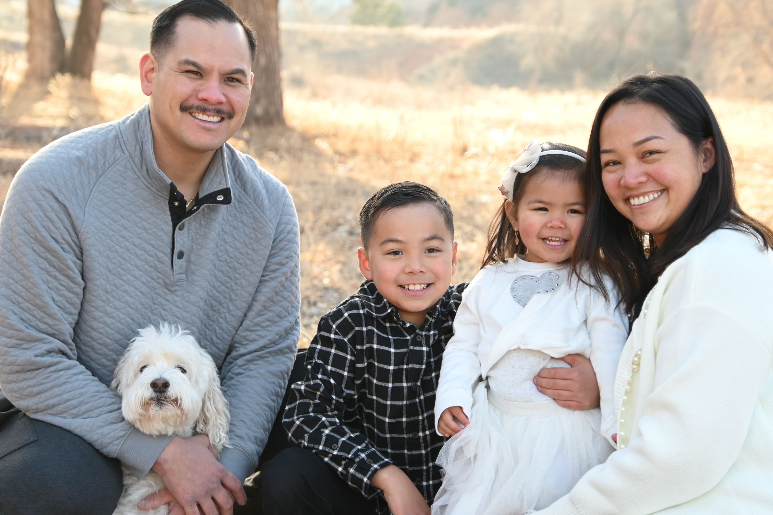 Family and small white dog pose together outdoors in coordinating casual outfits during a group photo session.