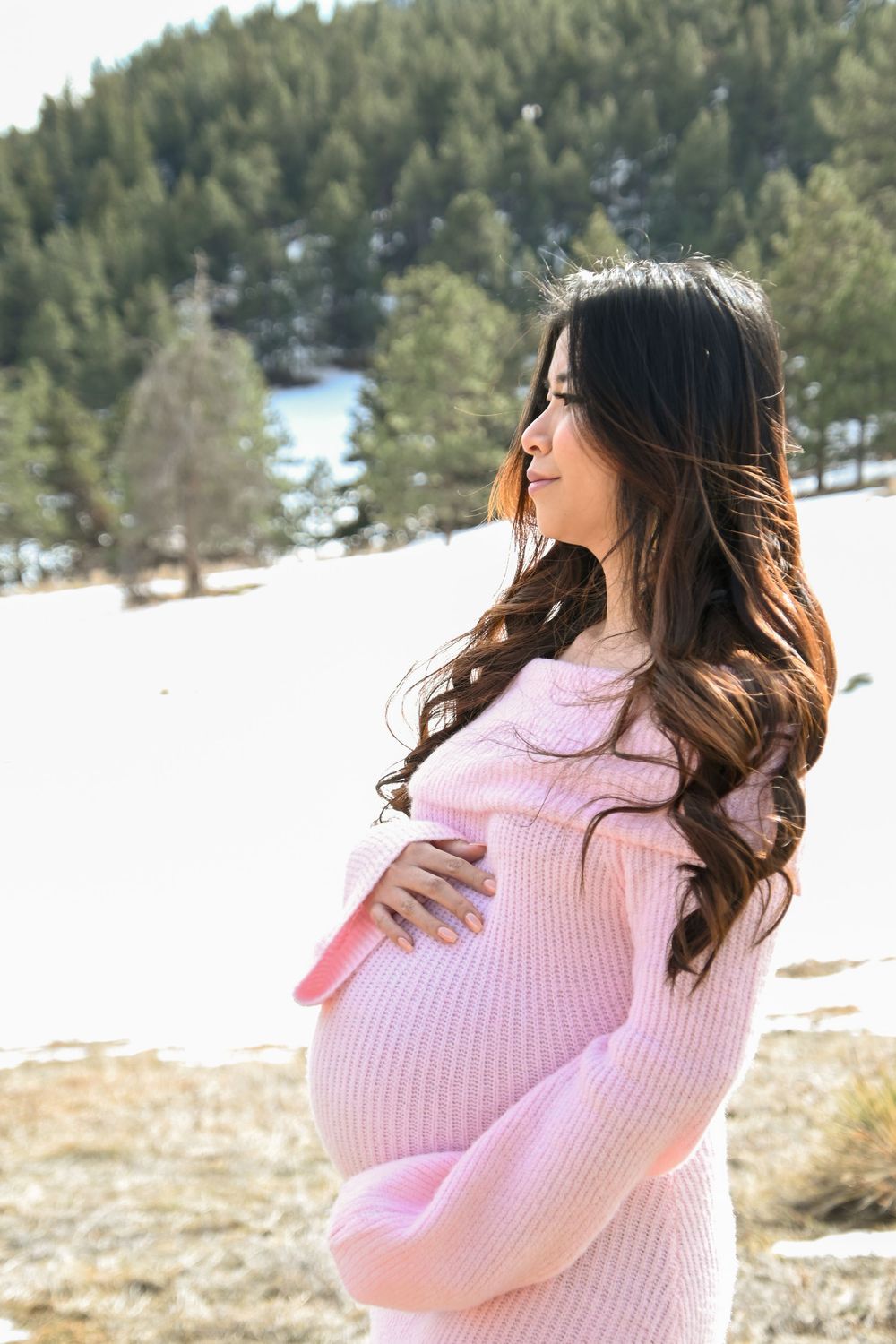 Expectant mother in pink sweater stands in snowy mountain landscape with pine trees in background.