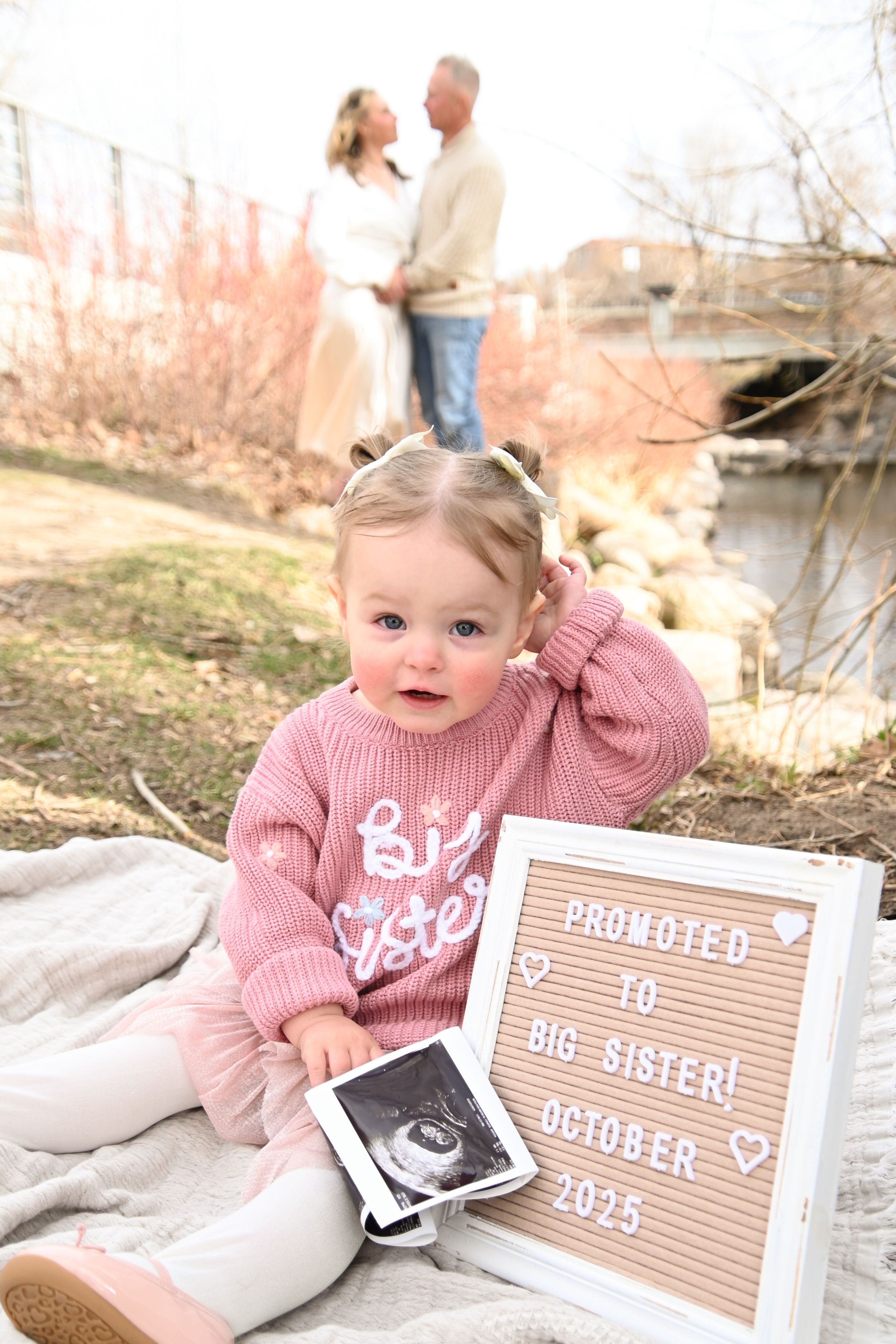 A baby in a pink sweater lies on a blanket next to letterboard signs making a pregnancy announcement for 2025.
