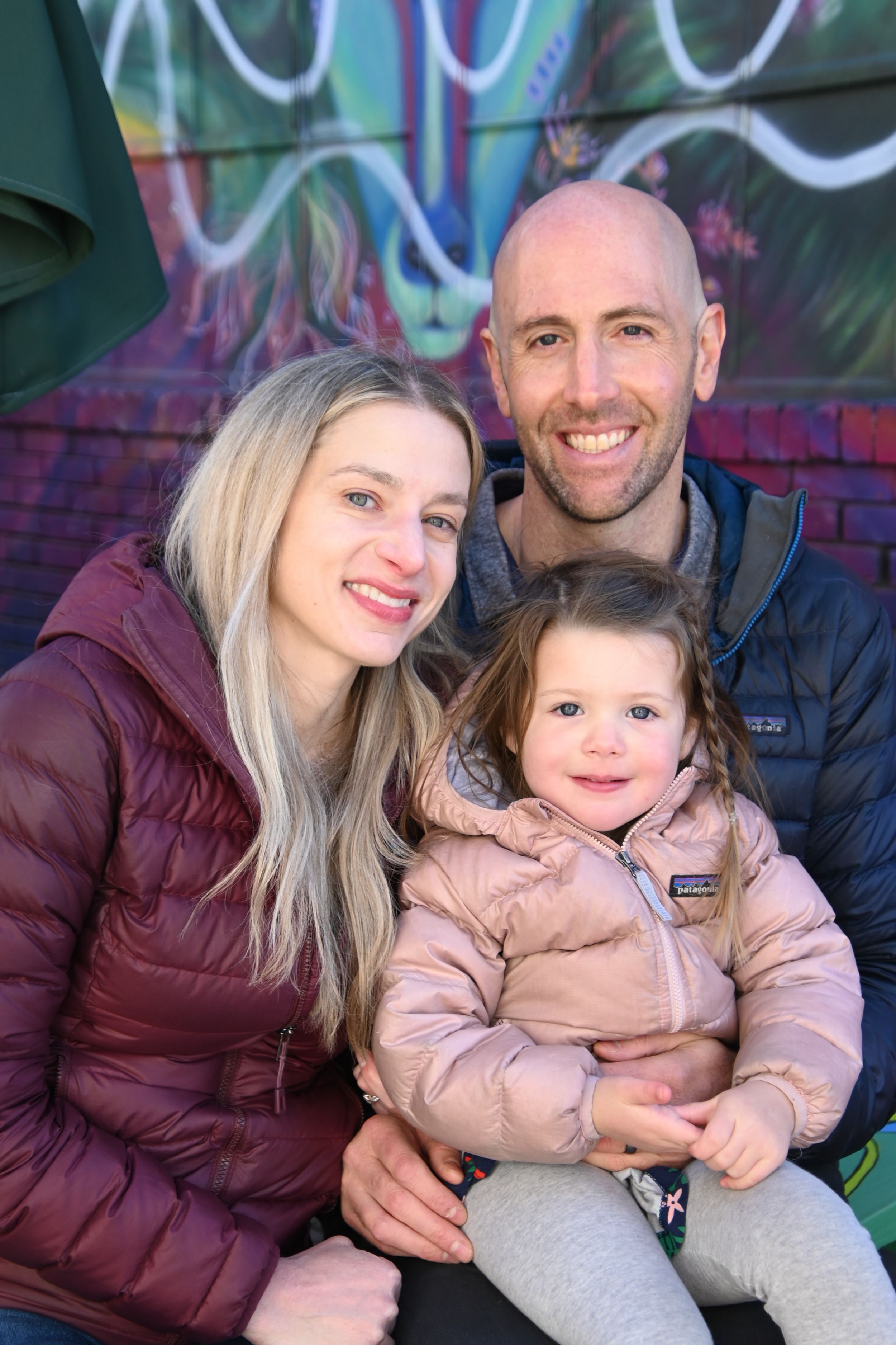 Family wearing warm winter coats poses together against colorful graffiti wall in outdoor photo series.