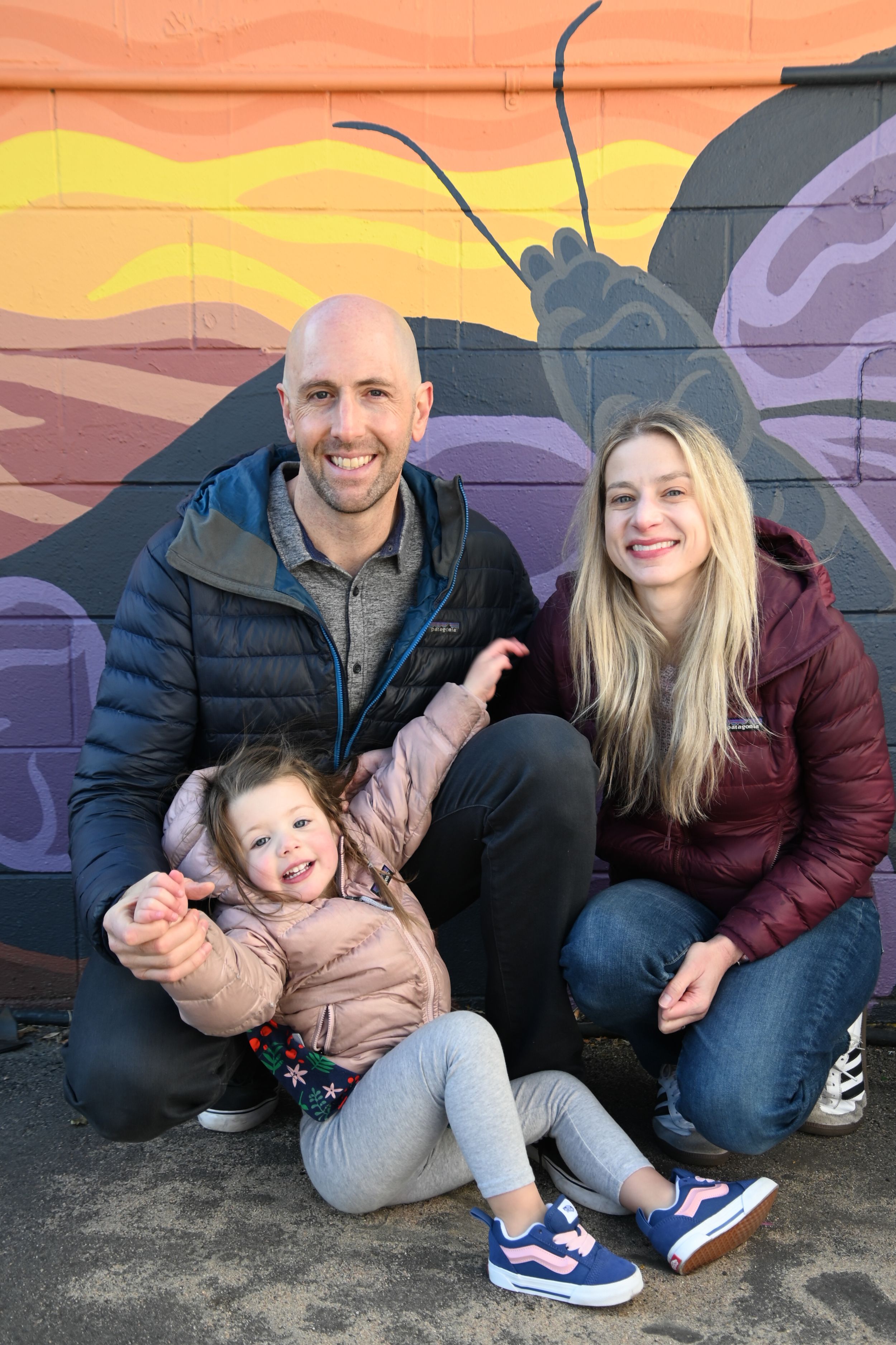 A family poses together in front of a colorful mural wall with abstract designs and a large snail at sunset.
