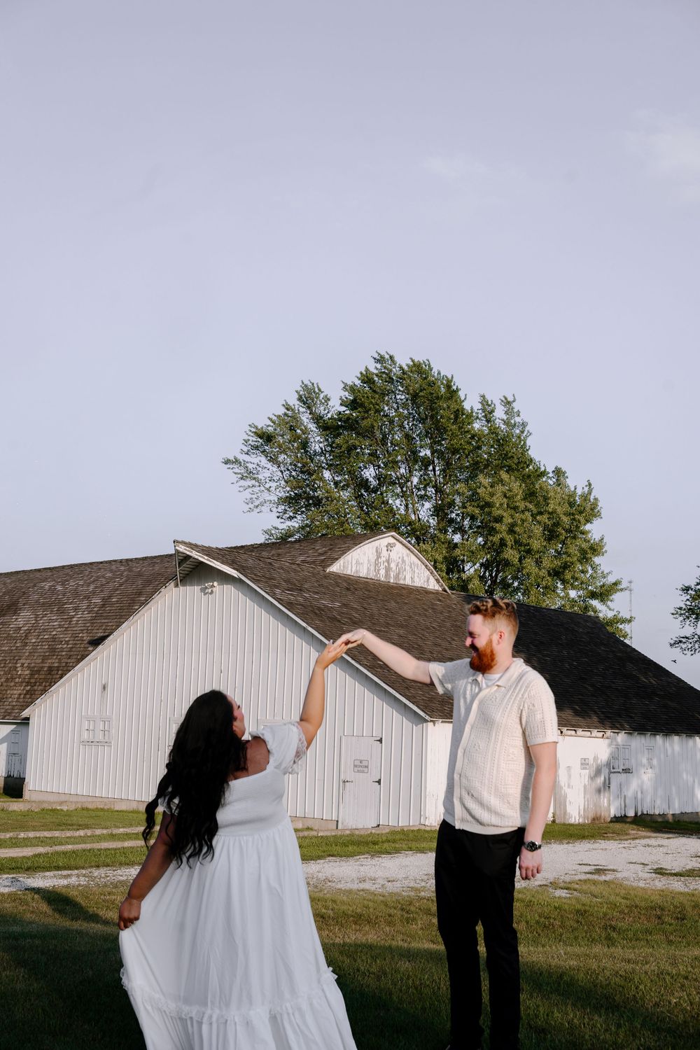 A couple dances playfully in a field with white barns in the background during a photo session.