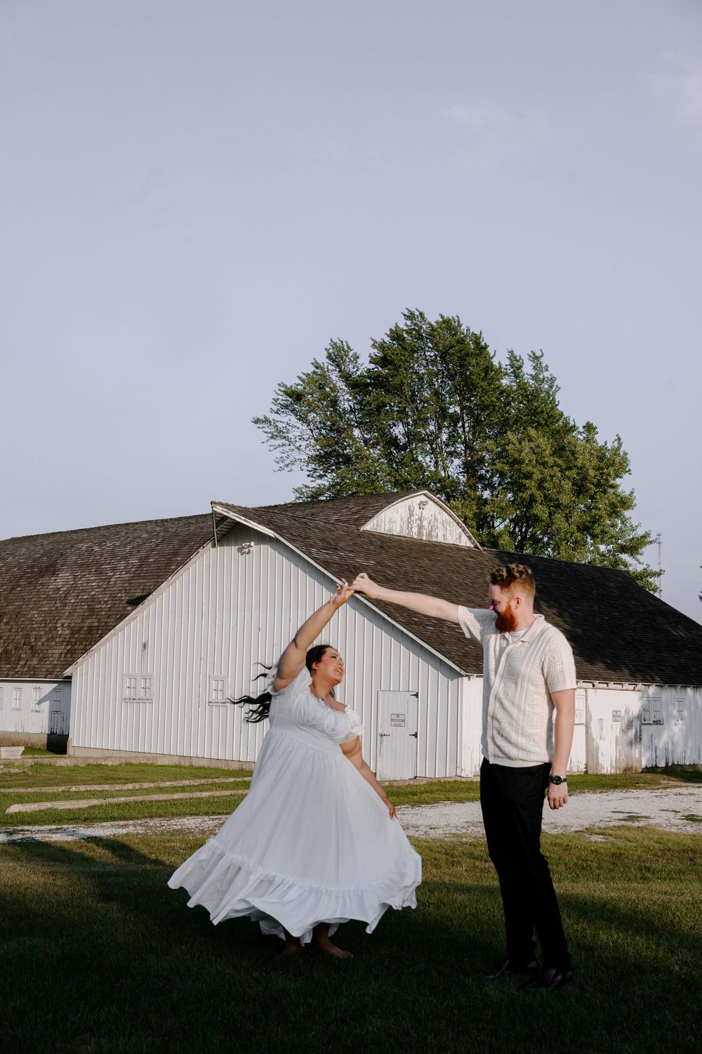 A couple twirls and dances together outside of a rustic white barn building at sunset.