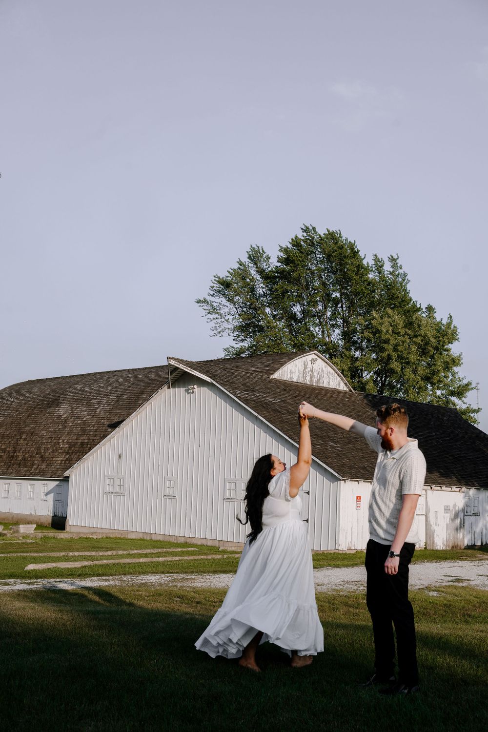 A couple dances together in front of a rustic white barn during a countryside photo session.