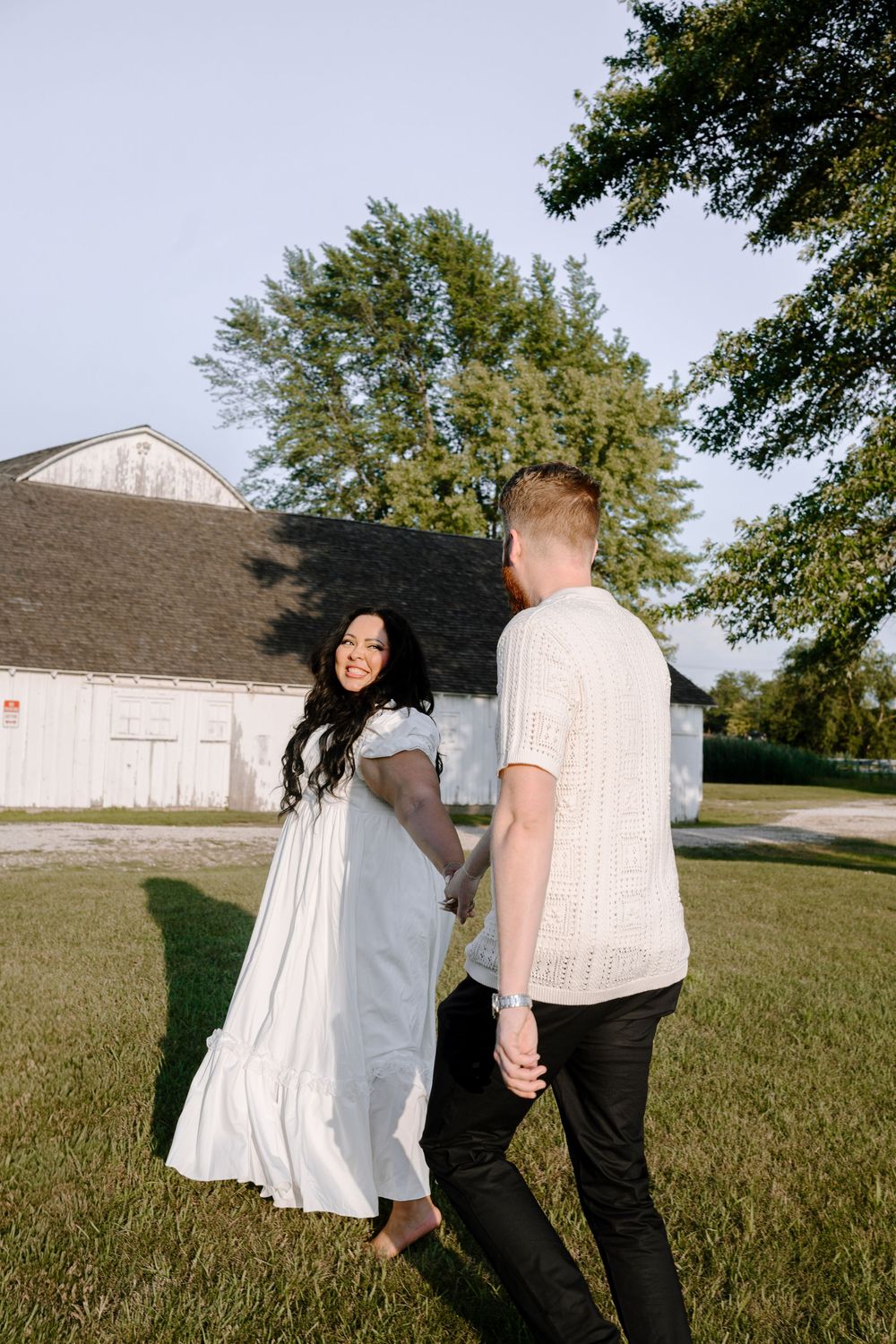 A couple enjoying a playful moment together on grass near a rustic white barn on a sunny day.