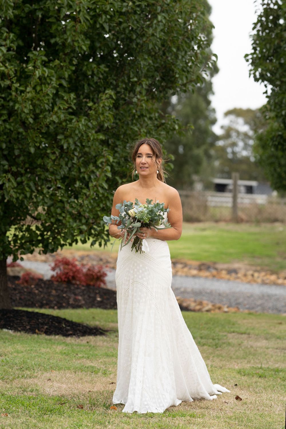 A bride in a strapless lace wedding dress holds a bouquet while standing in a garden setting with green trees.