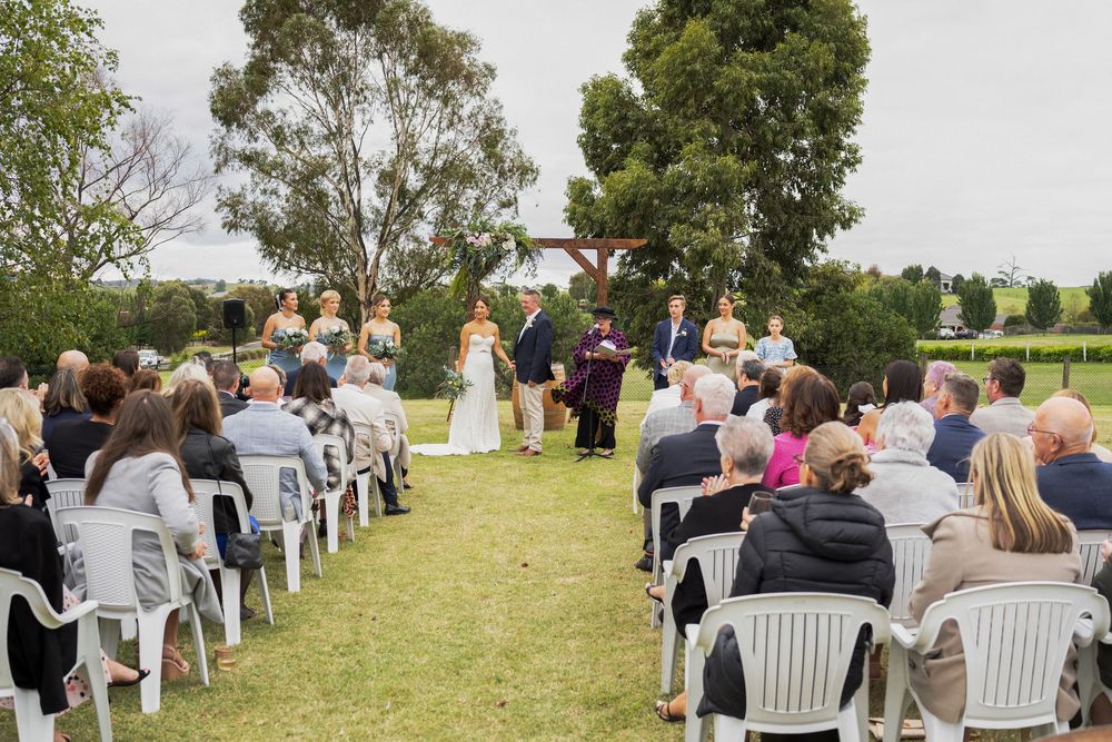 An outdoor wedding ceremony setup with white chairs on grass lawn surrounded by trees and guests in attendance.