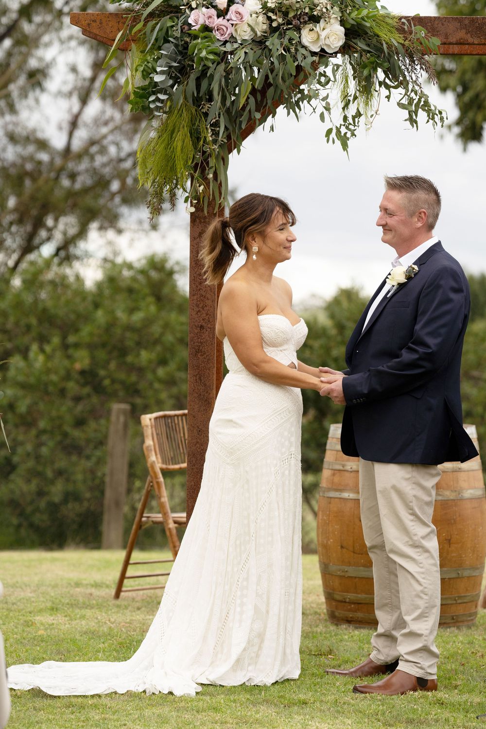 A couple stands at a rustic wedding ceremony arch decorated with greenery and flowers, with wine barrels nearby.