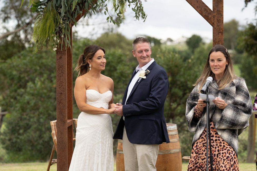 Three people stand under a rustic wooden arch during an outdoor wedding ceremony.