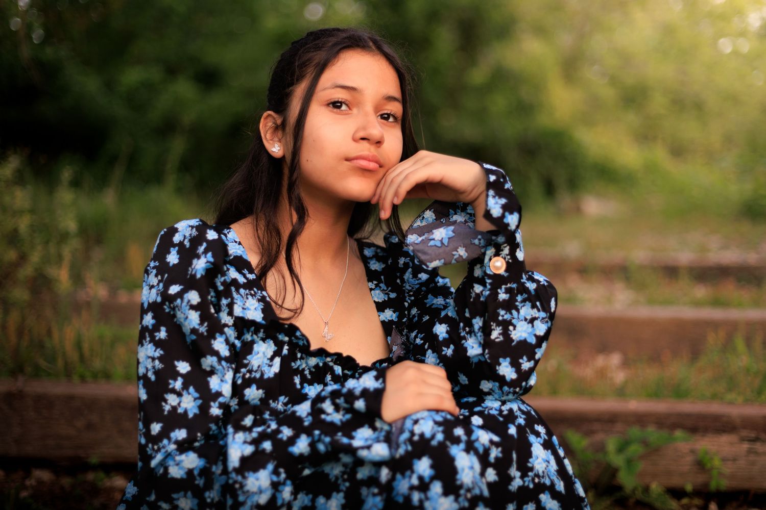 A person in a blue floral dress poses thoughtfully in a natural outdoor setting during golden hour lighting. In Pearland, TX