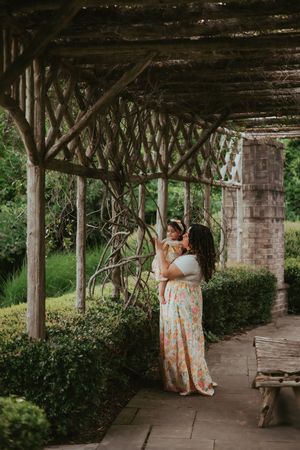 A mom and daughter in matching floral dresses admire the vines of the pergola they are in.