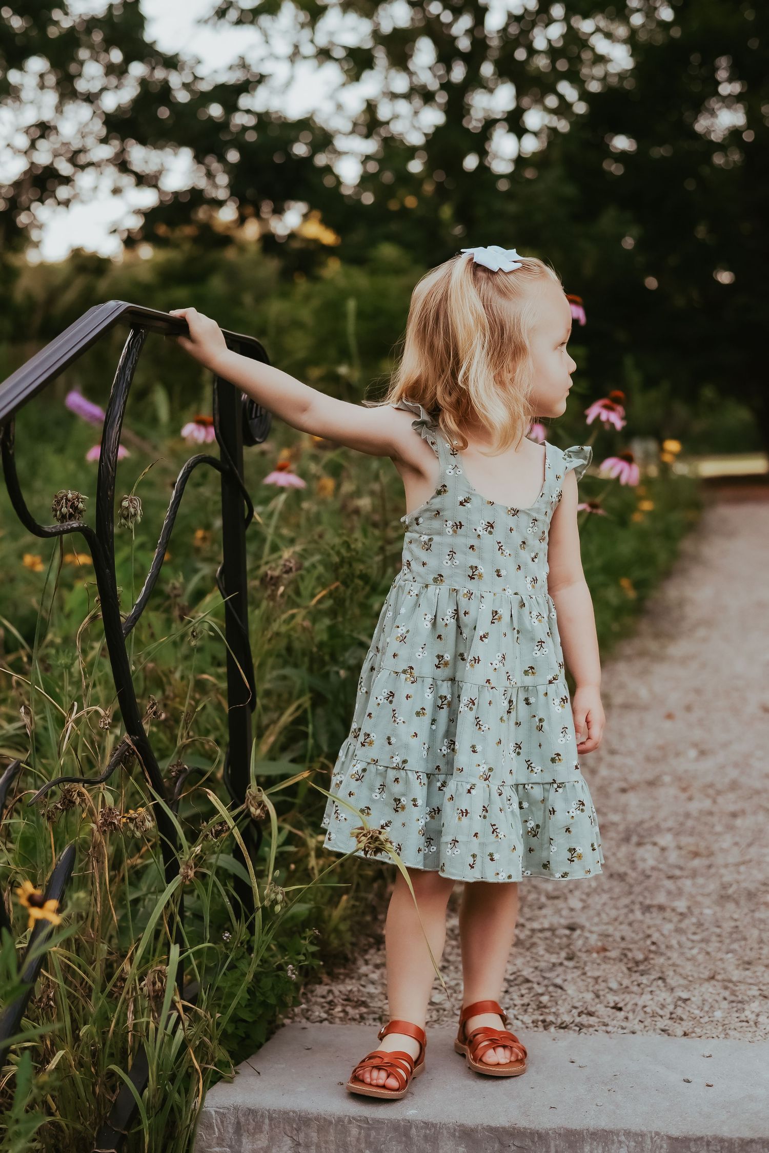 Young child in floral dress touches railing along garden path.