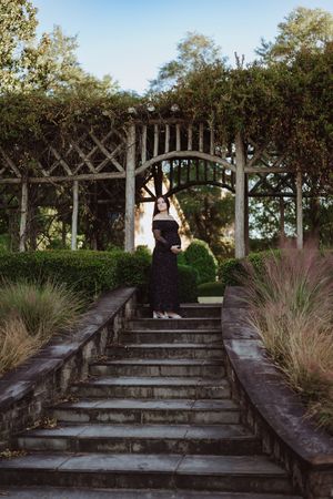 Woman in black dress stands on garden steps beneath a vine-covered pergola arch surrounded by lush greenery.