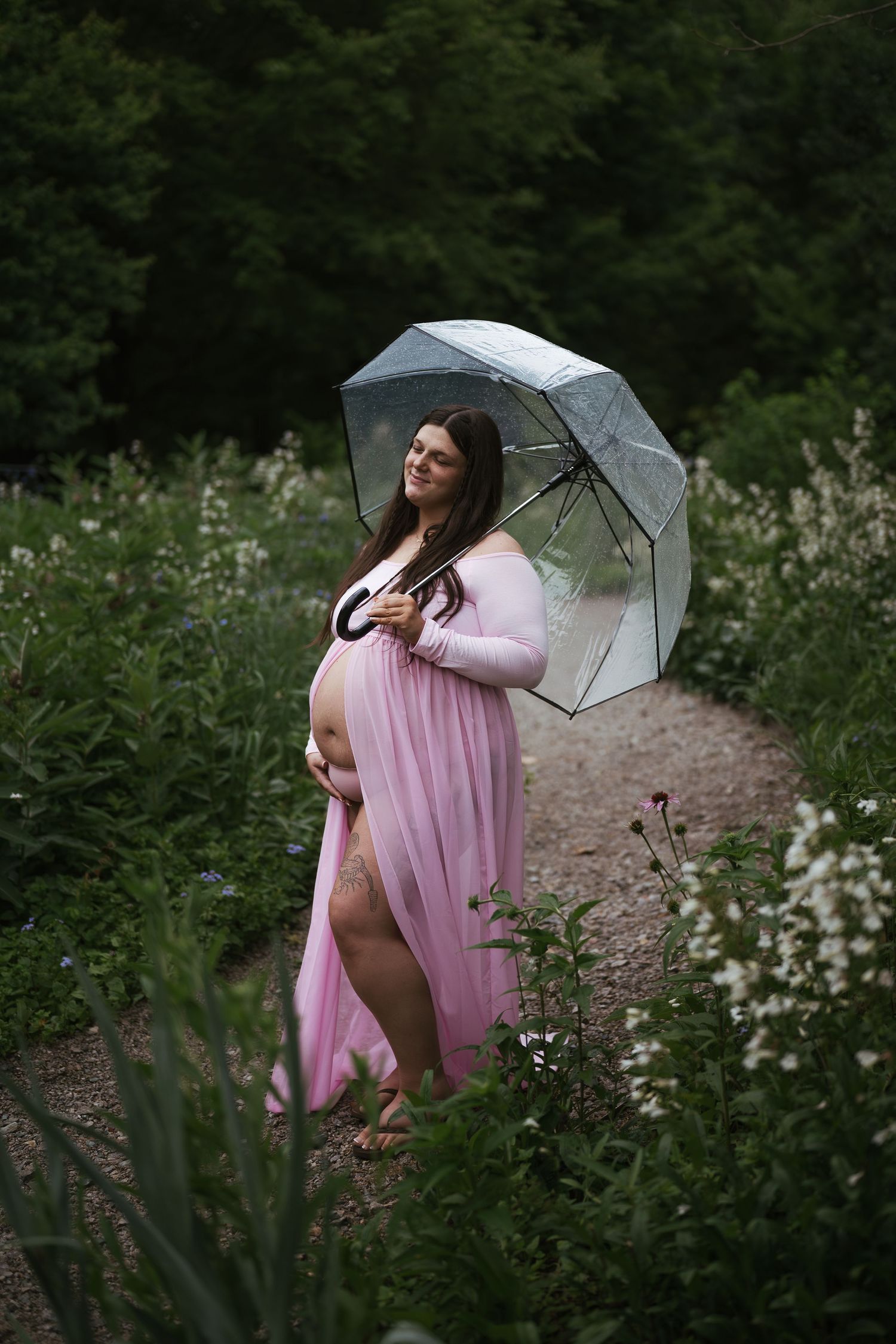 Pregnant woman in pink maternity gown holding clear umbrella on garden path surrounded by lush greenery.
