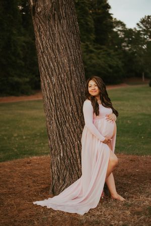 An expectant woman in a flowing pink dress cradles her baby bump while she poses against large tree trunk.