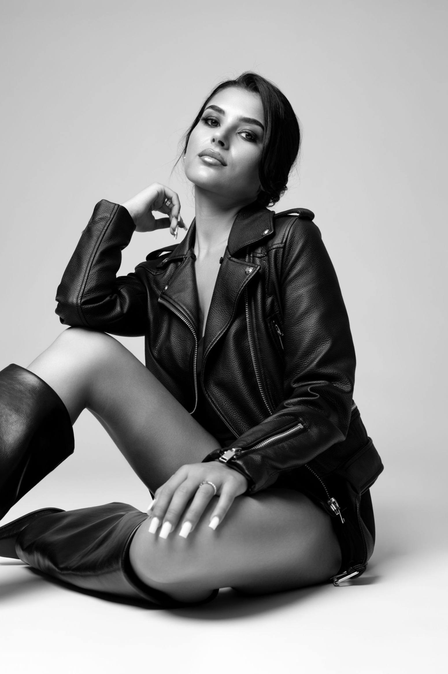 Black and white fashion photo showing model in leather motorcycle jacket and boots posing on studio floor.