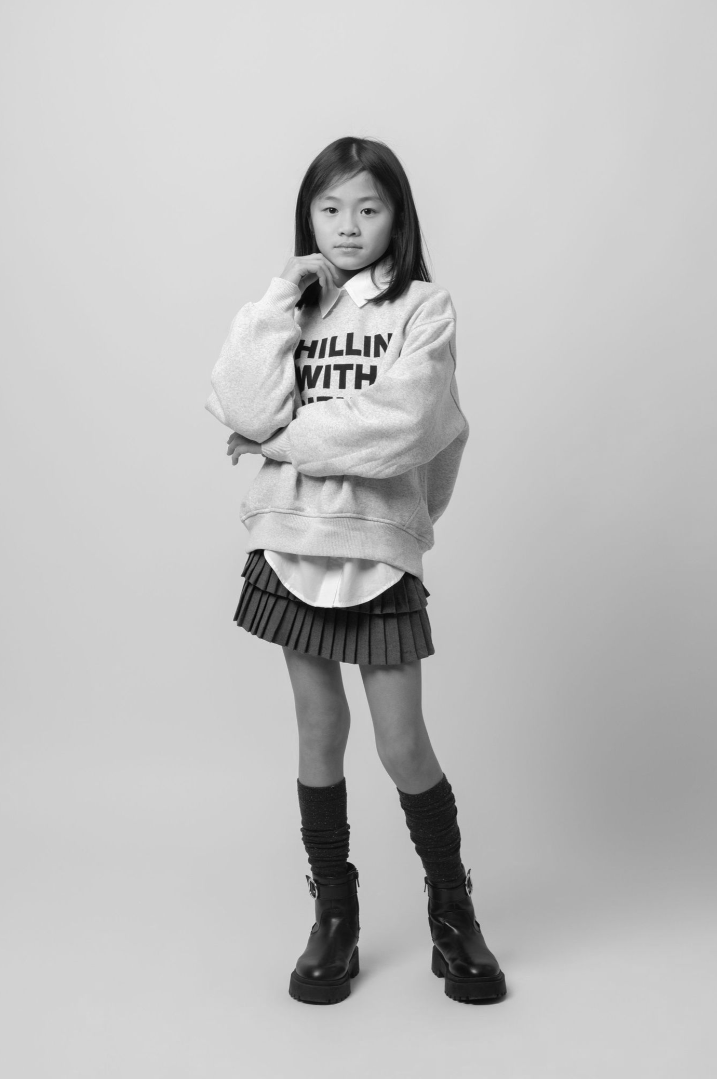 Black and white studio portrait showing someone in a white sweatshirt, pleated skirt and tall boots against a plain backdrop.