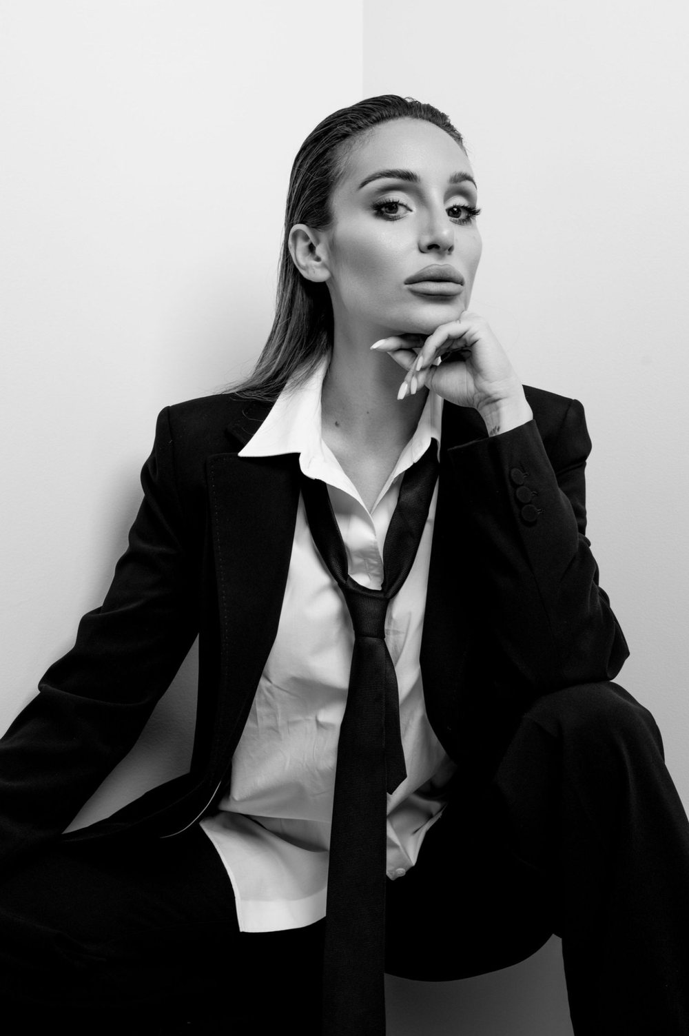 Black and white fashion portrait of someone in a sleek business suit with androgynous styling, sitting contemplatively.