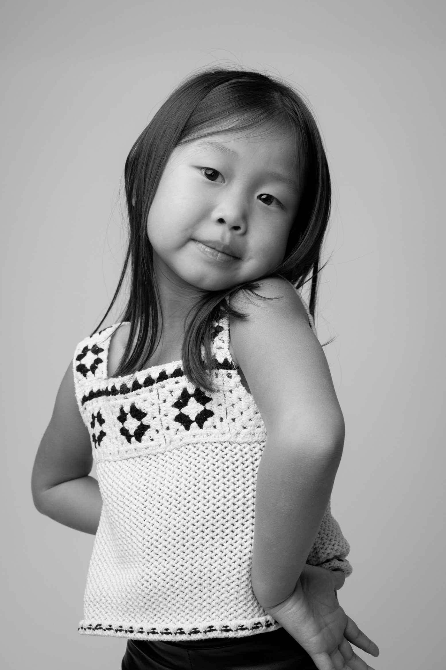Black and white portrait of a young child wearing a white crochet top with geometric patterns posing playfully against gray background.