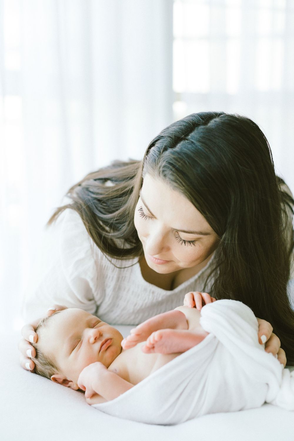 Tender moment between mother and sleeping infant on white bed.