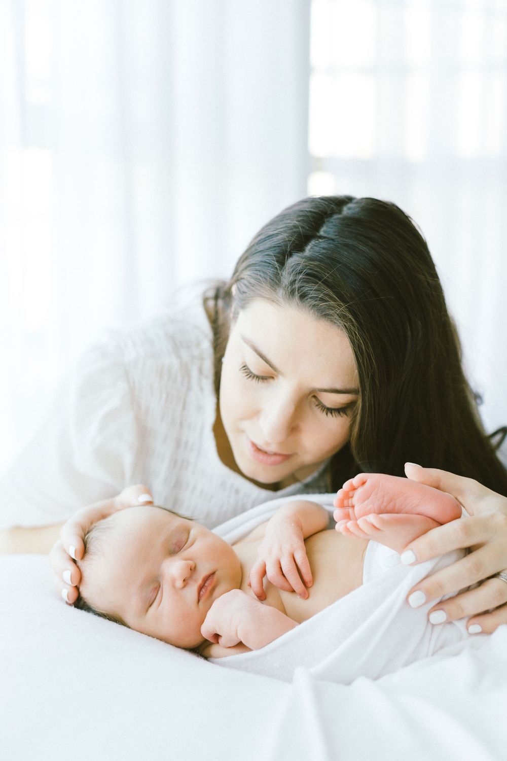 Parents lovingly gazing at their peaceful newborn baby in white bedding by a bright window.