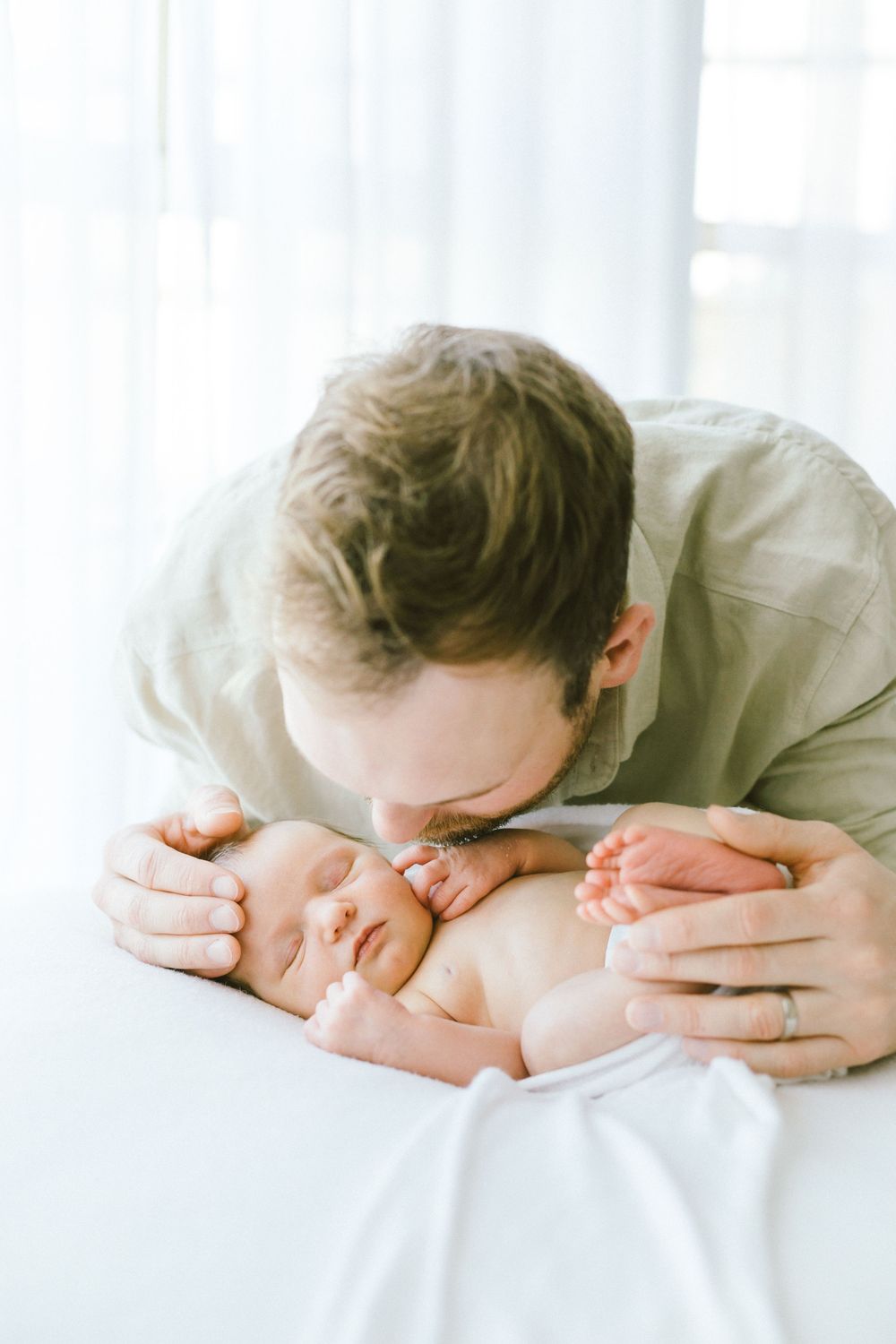 Parent tenderly kissing sleeping newborn baby on white sheets in natural light.