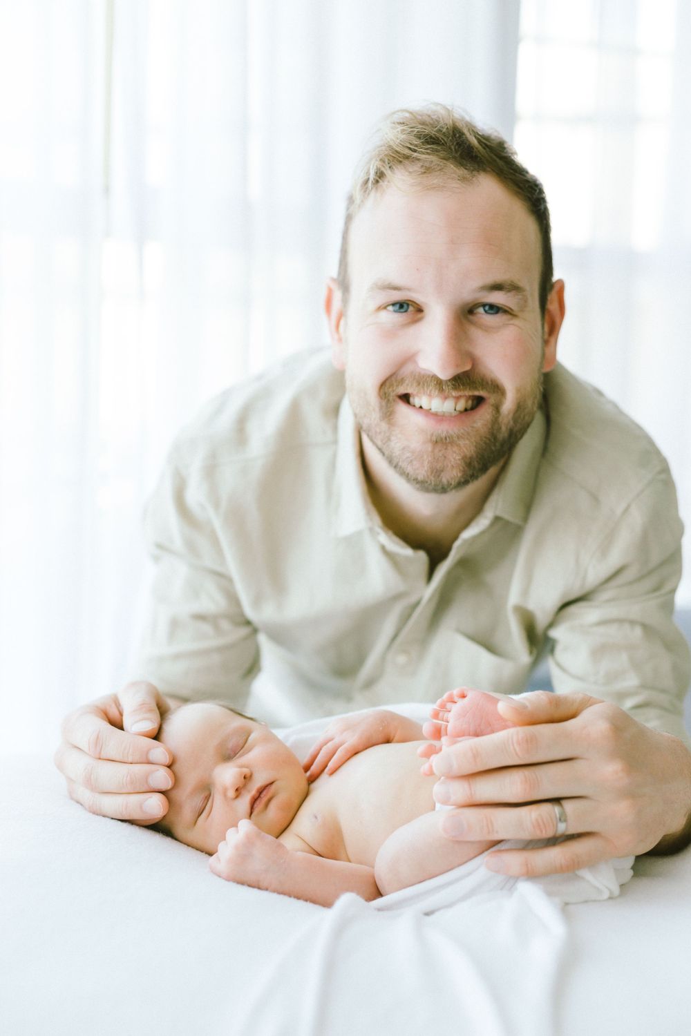 Parent smiling while bonding with their sleeping newborn on white bedding.