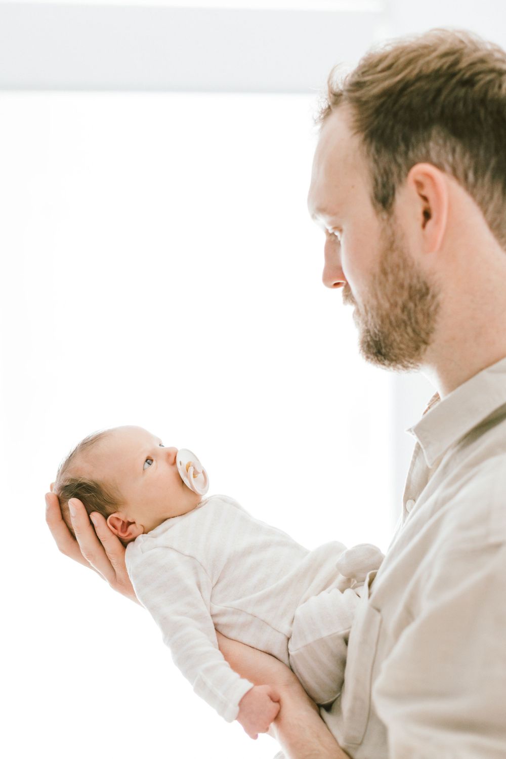 Parent in beige shirt tenderly holds newborn baby near bright window.