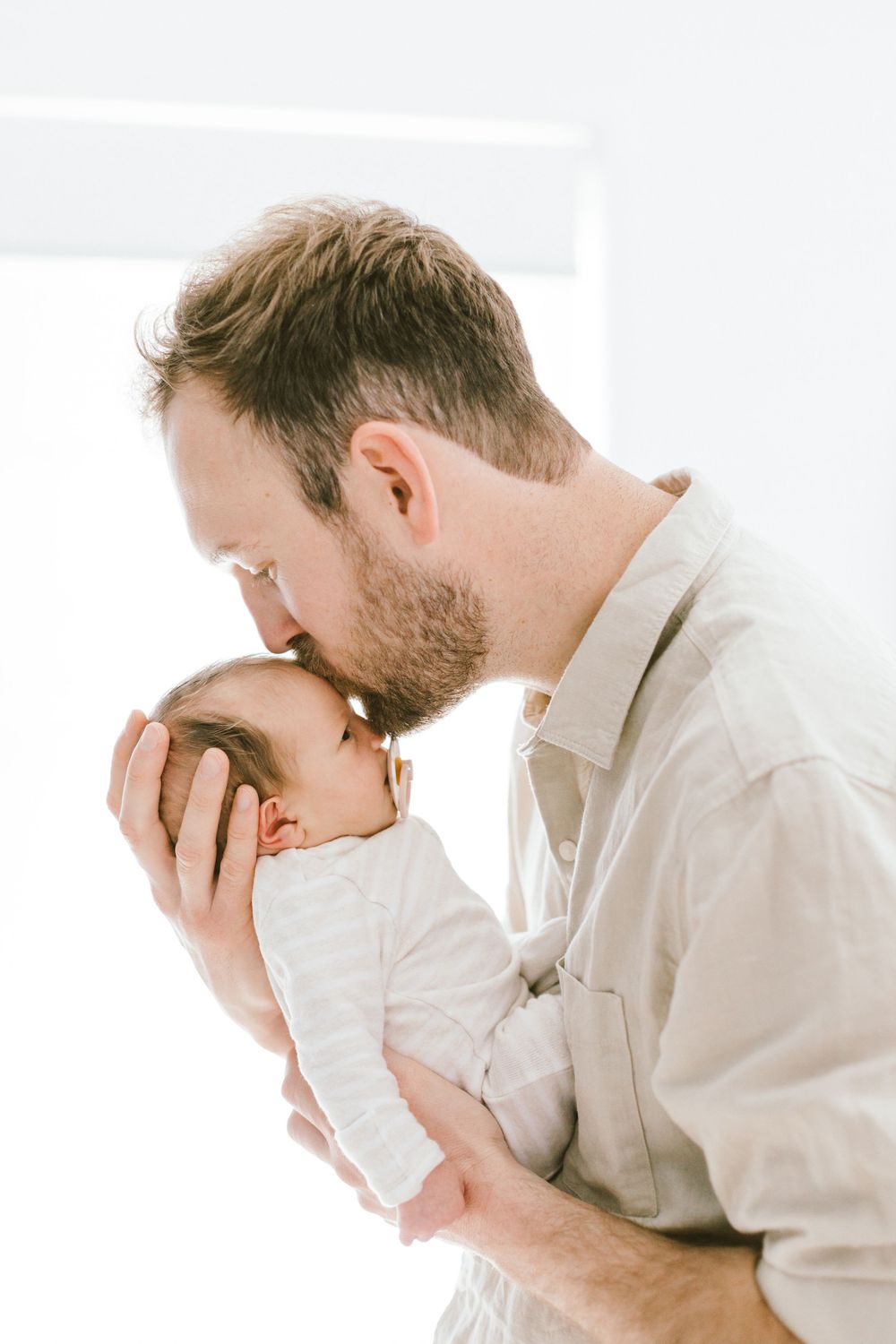 Parent in light colored shirt gives gentle kiss to newborn baby by window.