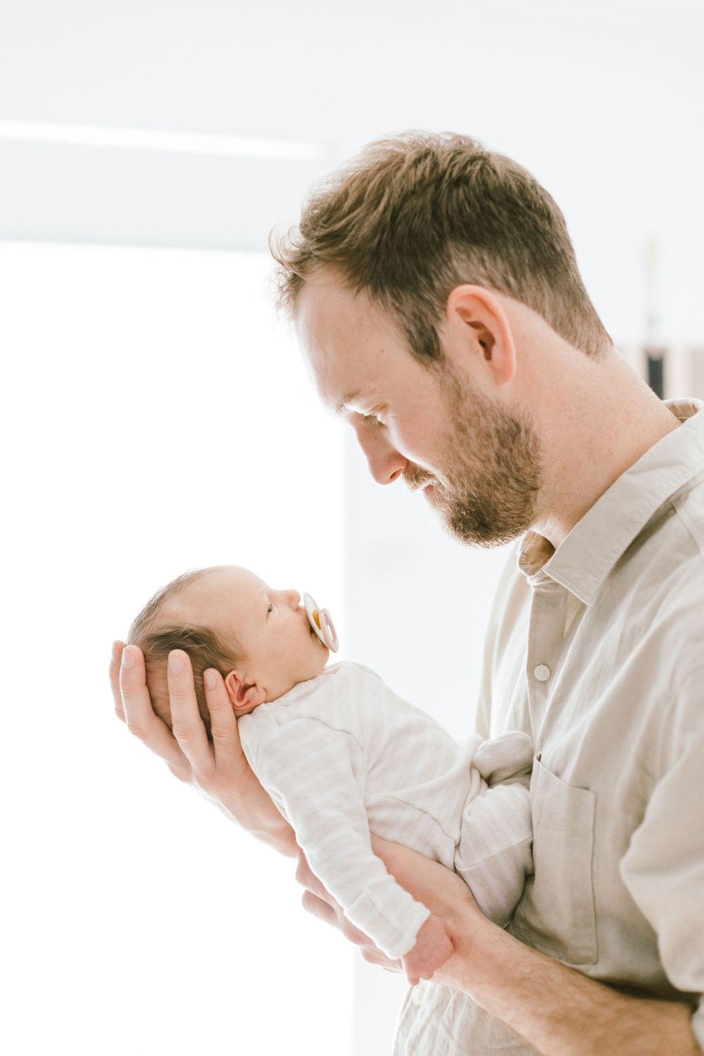 Tender moment between father and newborn baby in white against bright background.