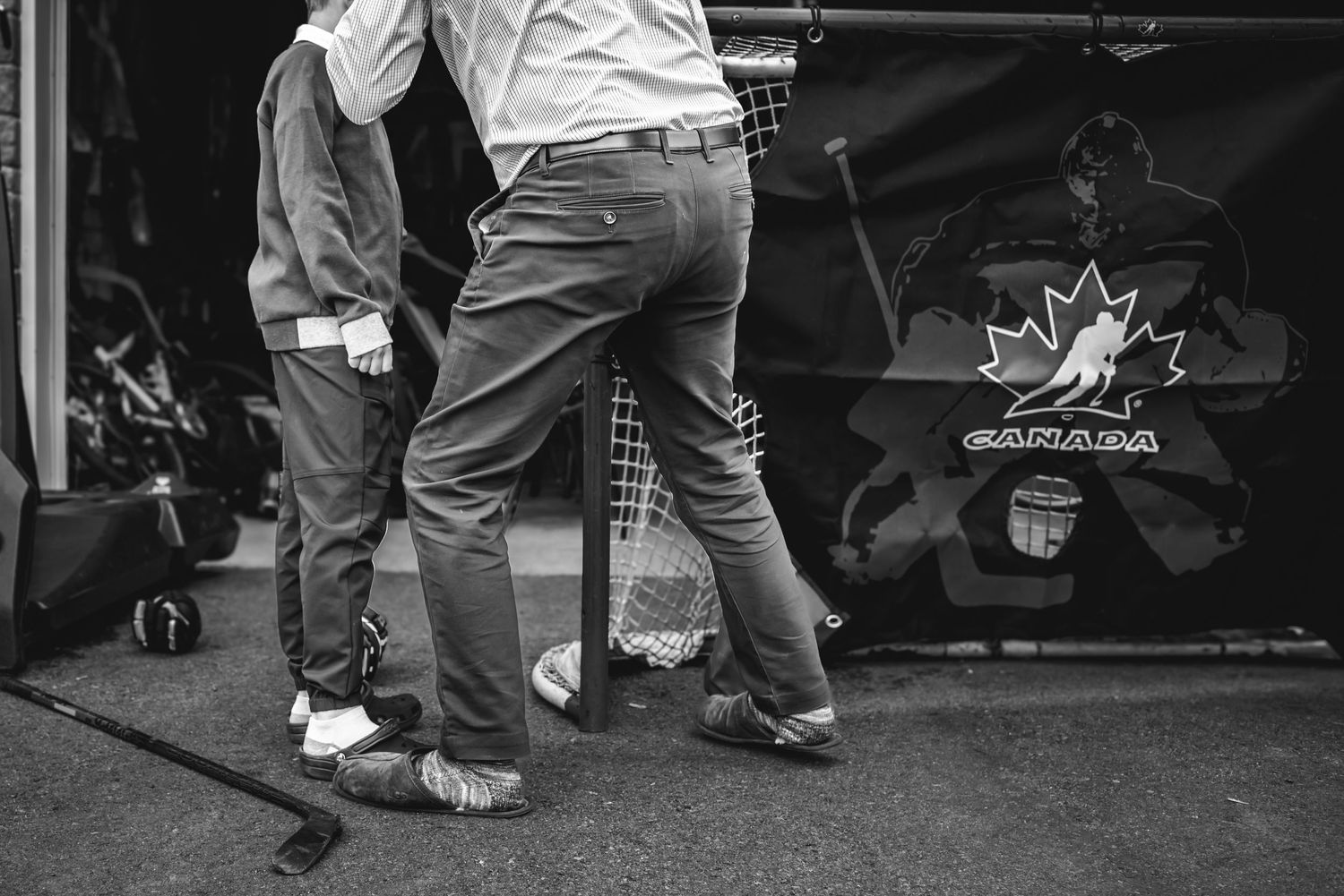 black and white image of the lower parts of 2 bodies standing in front of a hockey net, with a stick on the ground beside them.