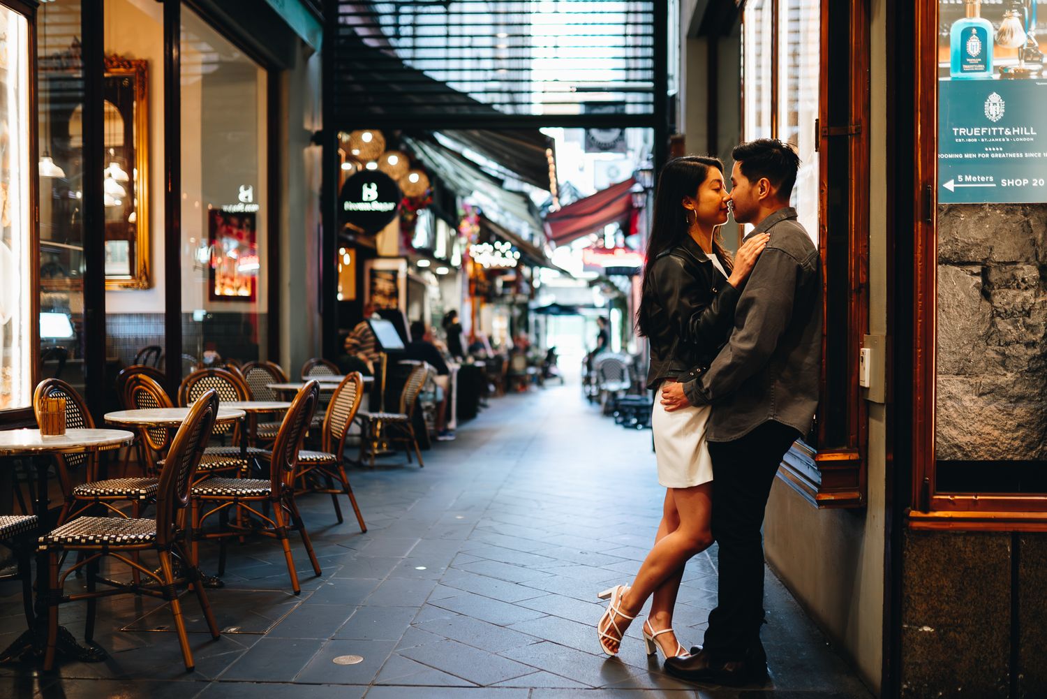 A couple embraces in an alley of Melbourne CBD, lined with cafe chairs and vintage storefronts taken by Melbourne Couples Photographer.
