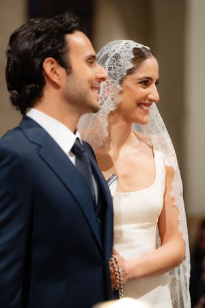 A bride in a white dress and lace veil stands next to her groom in a navy suit during their wedding ceremony.