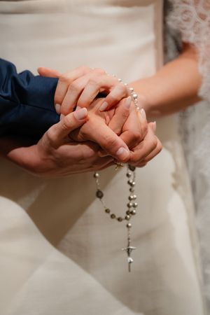 Close-up of hands holding rosary beads during wedding ceremony with white dress and navy suit visible in frame.