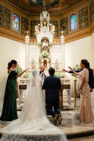 Bride and groom stand at ornate church altar during traditional Catholic wedding ceremony with veil flowing behind.