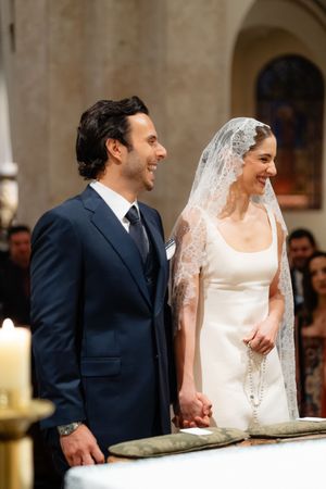 A joyful bride in a white dress and lace veil stands beside her groom in a navy suit during a church wedding ceremony.