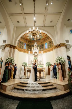 A wedding ceremony takes place in an elegant church sanctuary with ornate decor, chandeliers and floral arrangements.