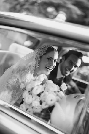 Black and white photo of a joyful newlywed couple in a car, with the bride holding a bouquet of white flowers.