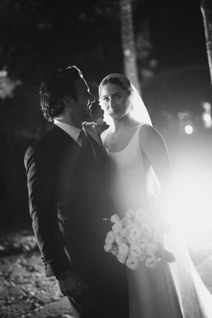 Atmospheric black and white wedding portrait showing couple in silhouette with veil and bouquet backlit by dramatic lighting.