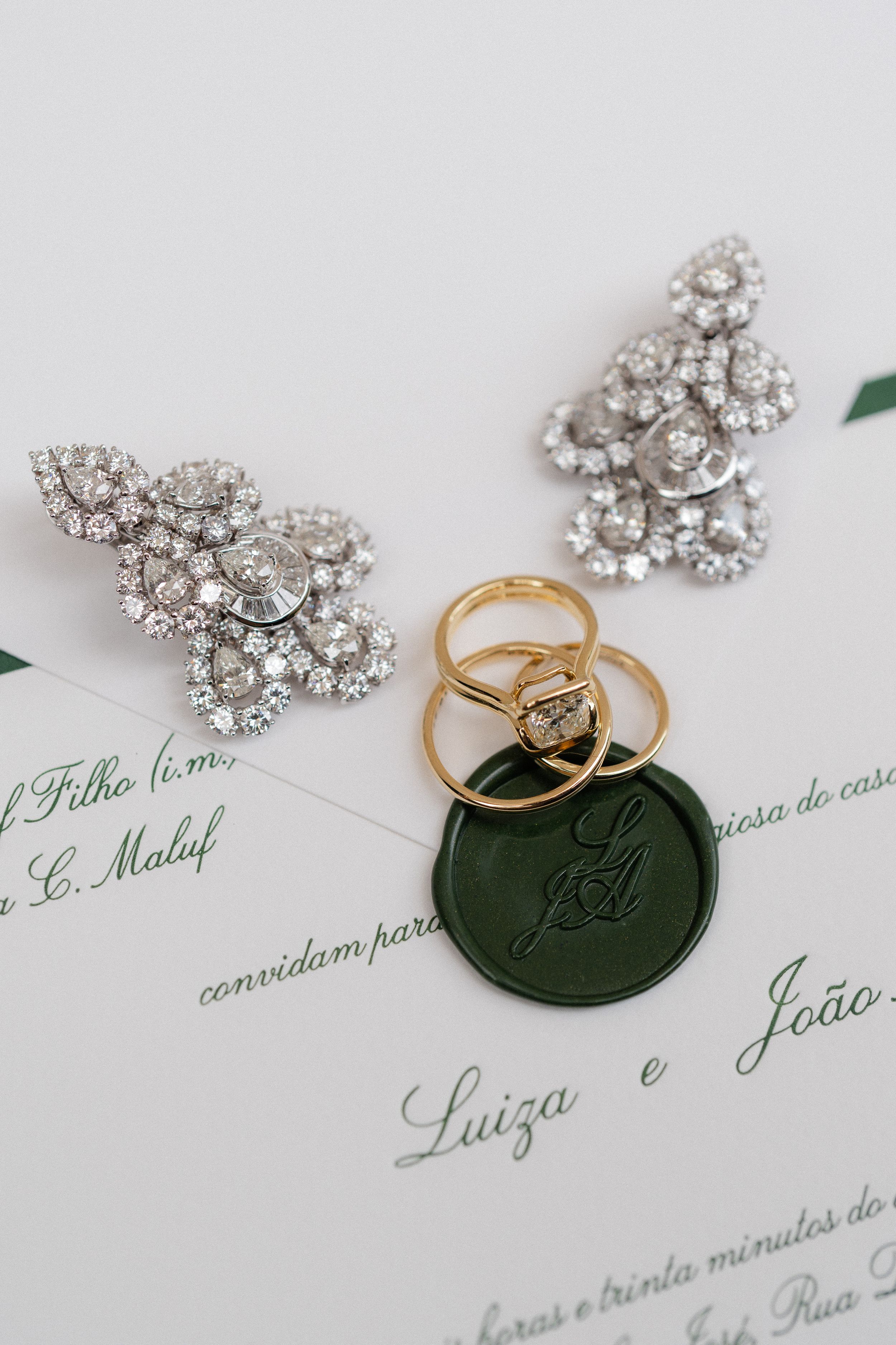 Wedding invitation with elegant diamond earrings, gold rings and a dark green wax seal on white paper.