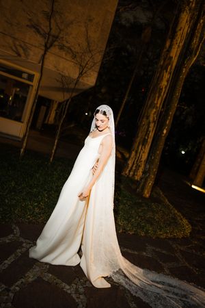 A bride in an elegant white wedding gown and flowing veil stands in a dimly lit outdoor setting at night.